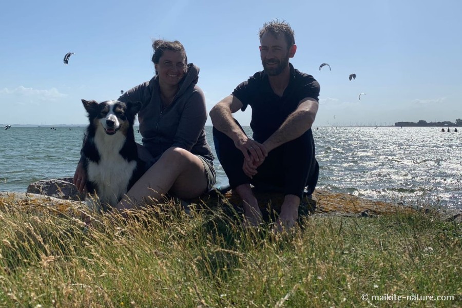 Ute, Daniel und Border Collie Luna sitzen an der Bucht vor Lemkenhafen auf Fehmarn, im Hintergrund Kitesurfer